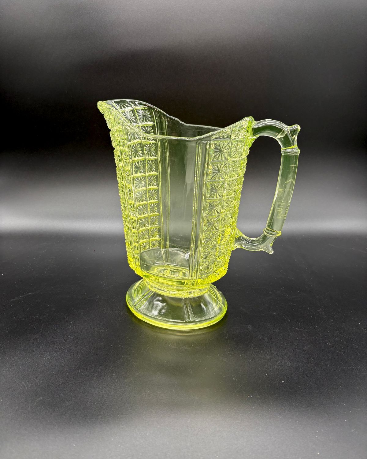Glowing canary yellow vaseline glass pitcher with a pressed daisy and button pattern, paneled body, fluted footed base, and a bold handle, set against a dark background.
