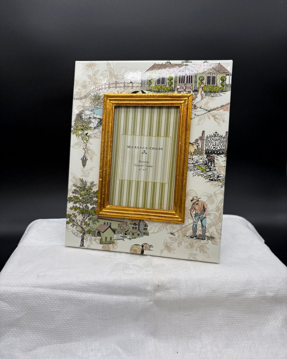 A retired MacKenzie-Childs Aurora enamel picture frame with a cream border hand-painted in a pastoral toile of cottages, trees, sheep, and a farmer, around a gold inner frame.