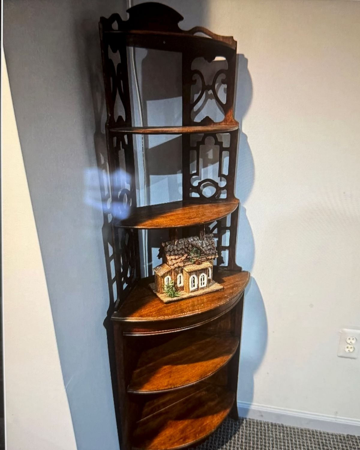 A tall dark wood corner etagere with three open display shelves, pierced fretwork side panels, a carved scalloped crest, and a bowfront lower cabinet, set in a room corner.