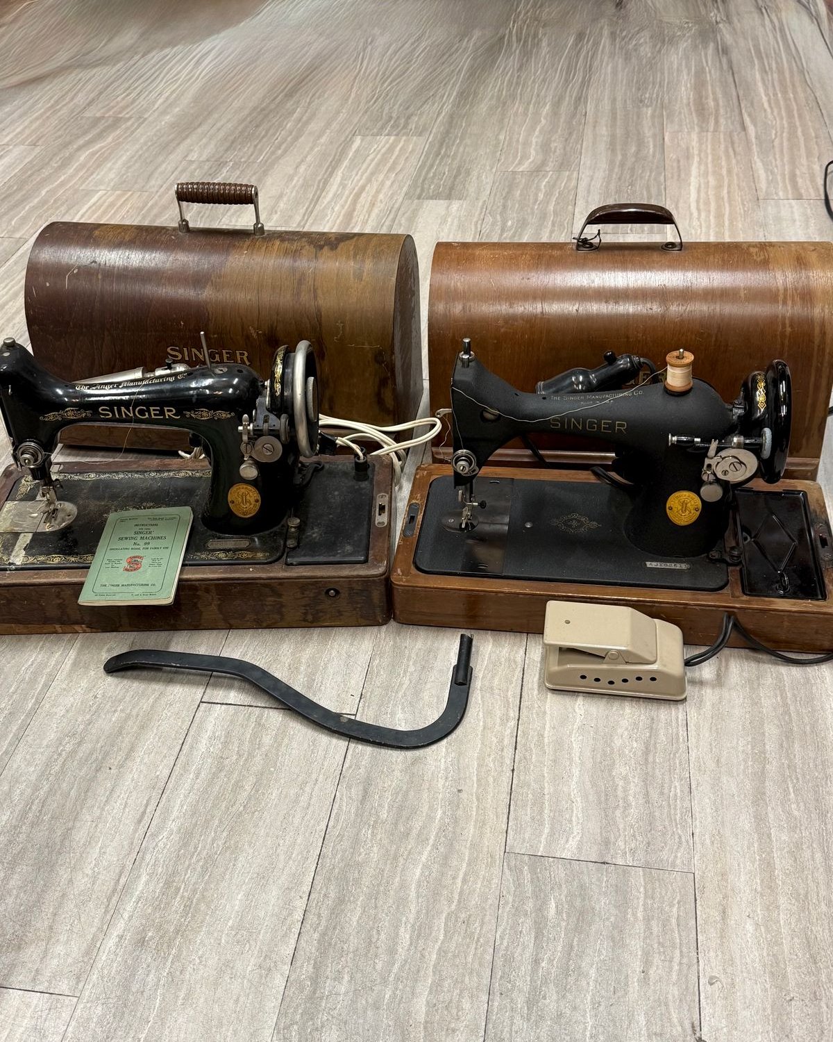 Two vintage black-and-gold Singer sewing machines side by side, each with a domed bentwood case; the 1927 on the left with its instruction book and curved knee bar, the 1949 on the right with a cream foot pedal.