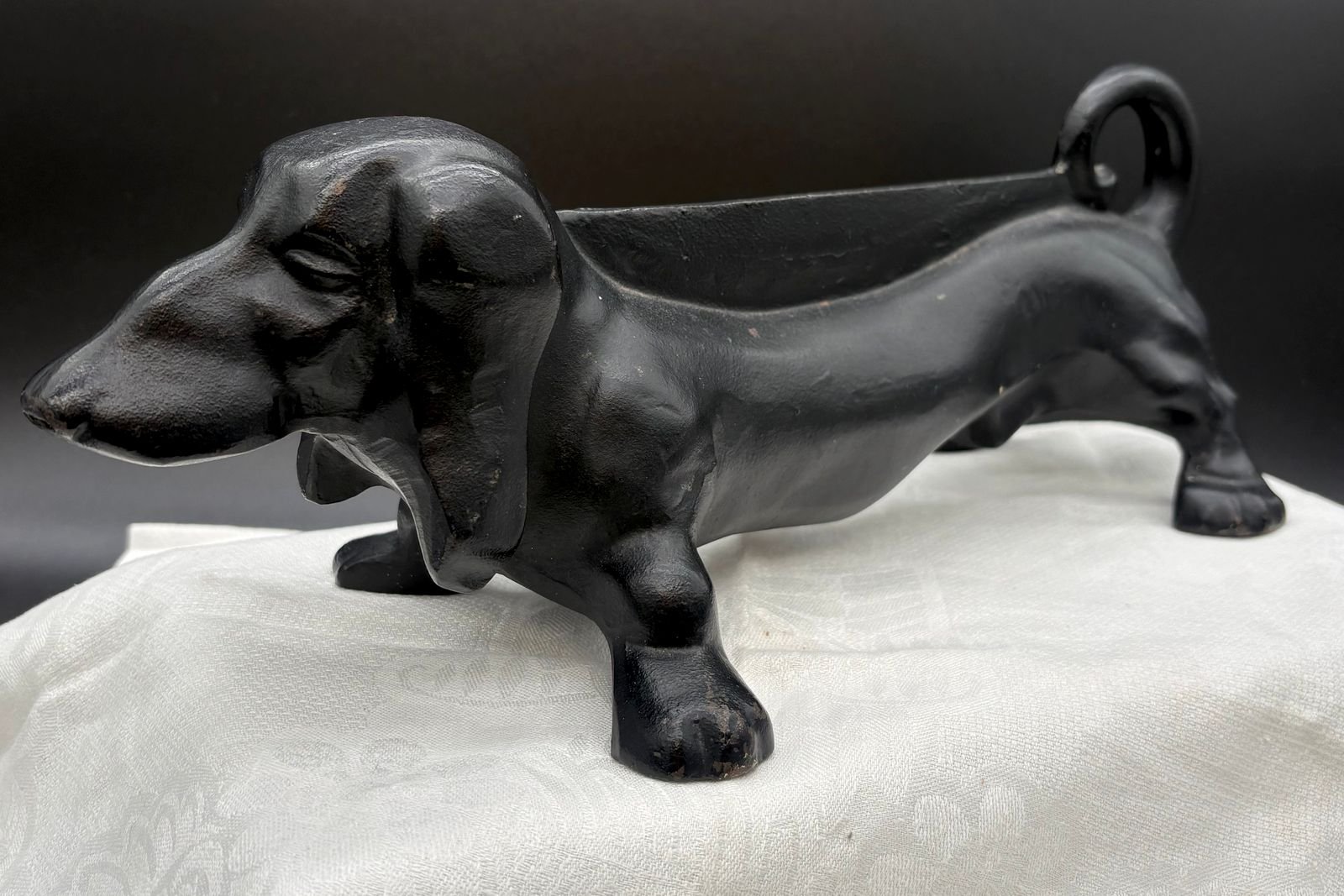 A black cast iron dachshund boot scraper and doorstop with floppy ears, a long arched back, four stout legs, and a curled tail handle, on a white cloth.