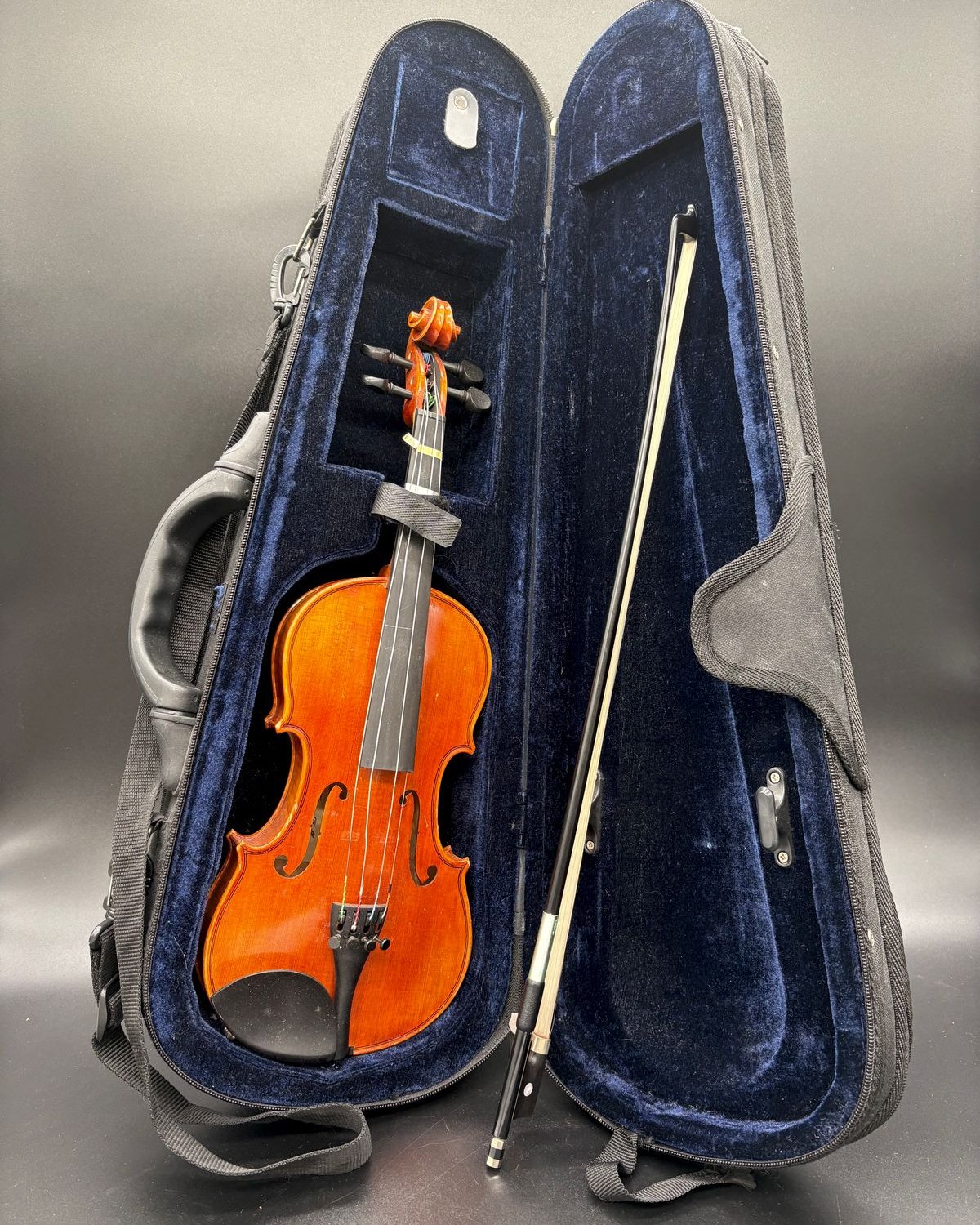 A child's Franz Hoffmann violin with reddish-brown varnish resting in an open black case lined with blue plush, with its bow in the lid.