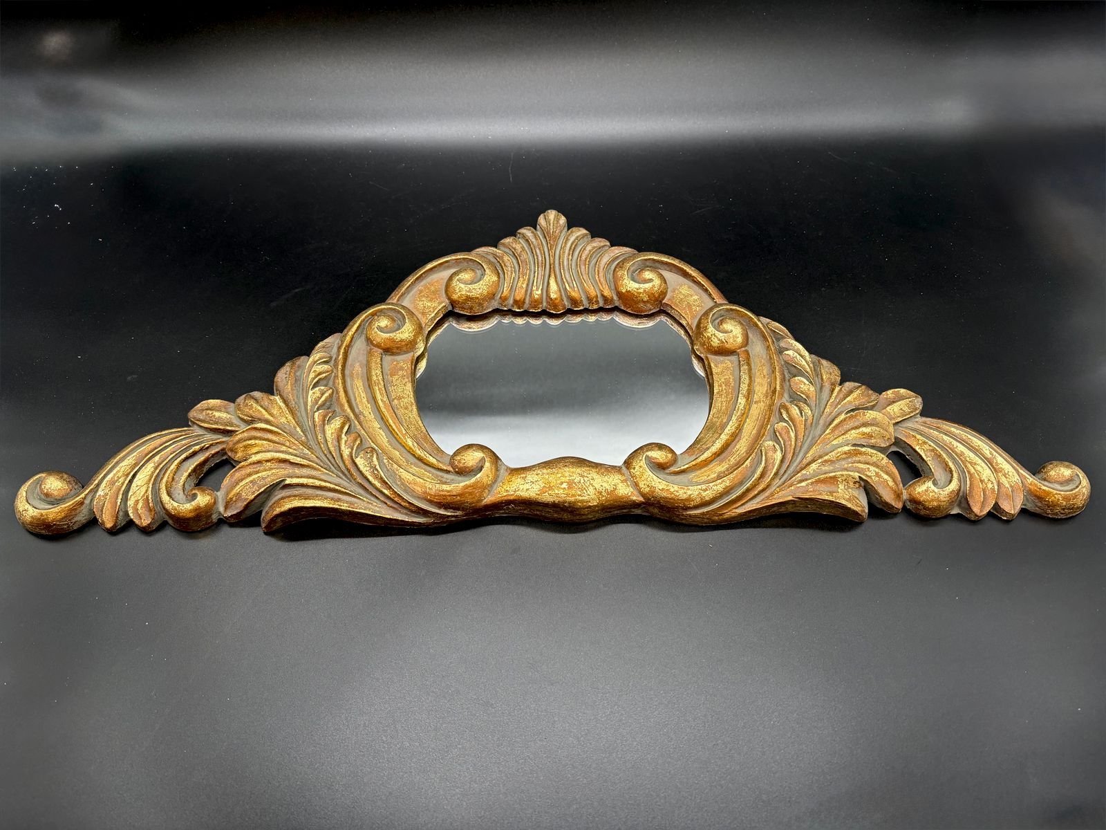 A wide carved giltwood wall mirror with Baroque and Rococo scrolls, curling acanthus leaves, and a shell crest framing a small central looking glass, on a dark background.