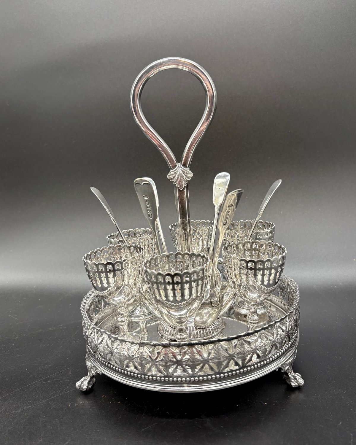 A Victorian silver-plated egg cruet: six pierced egg cups and six egg spoons arranged around a central looping handle on a round paw-footed stand.
