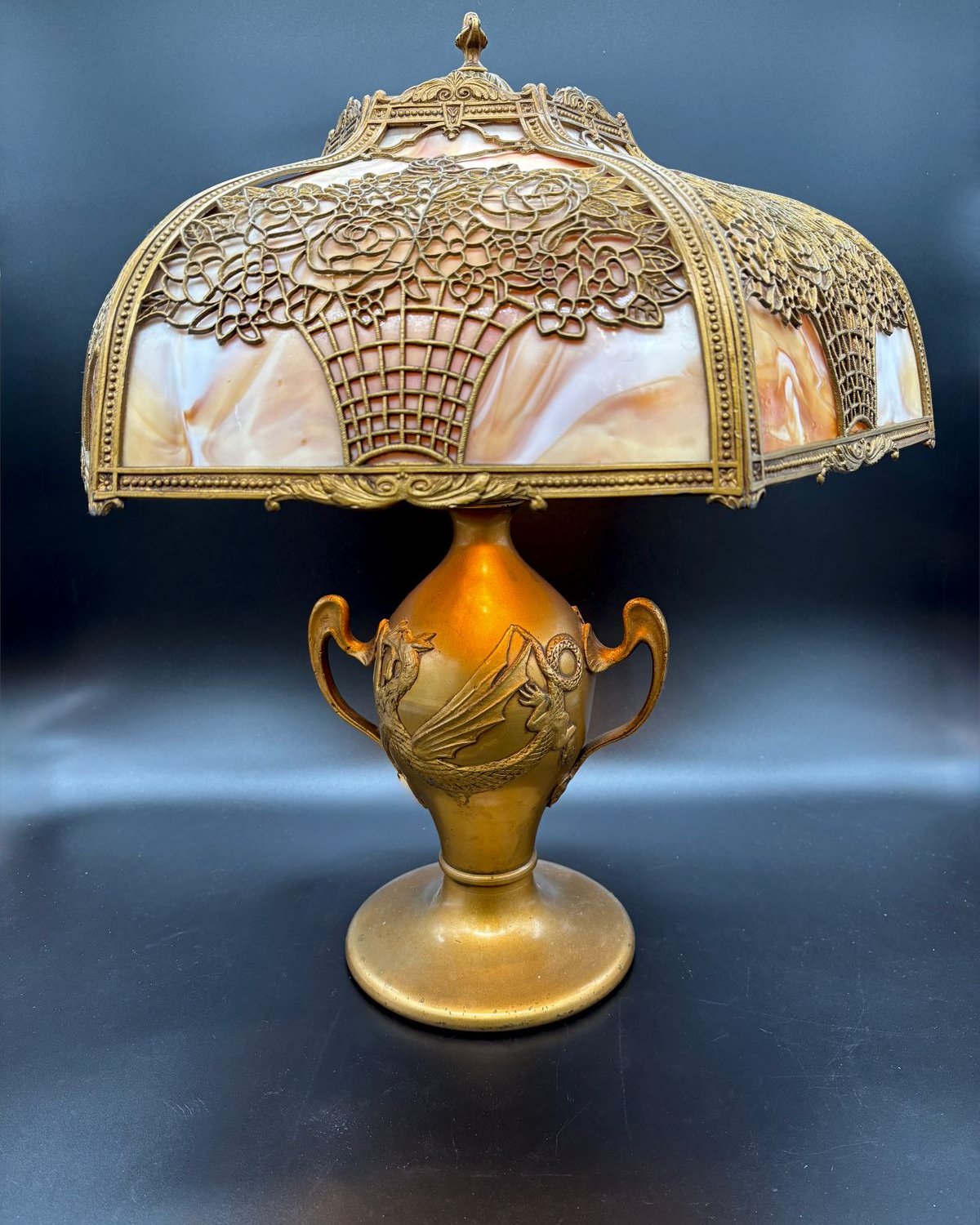 An Art Nouveau slag glass table lamp: a caramel glass shade under a gilt flower-basket filigree overlay, on a gilded urn base with a winged-dragon relief.