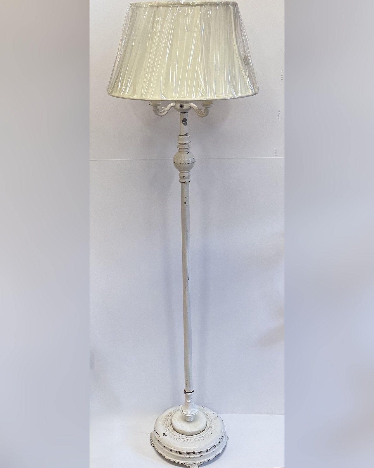 A tall antique floor lamp of white painted cast iron with a reeded column, urn-shaped knop, a decorative round base with chipped paint, and a cream pleated shade.