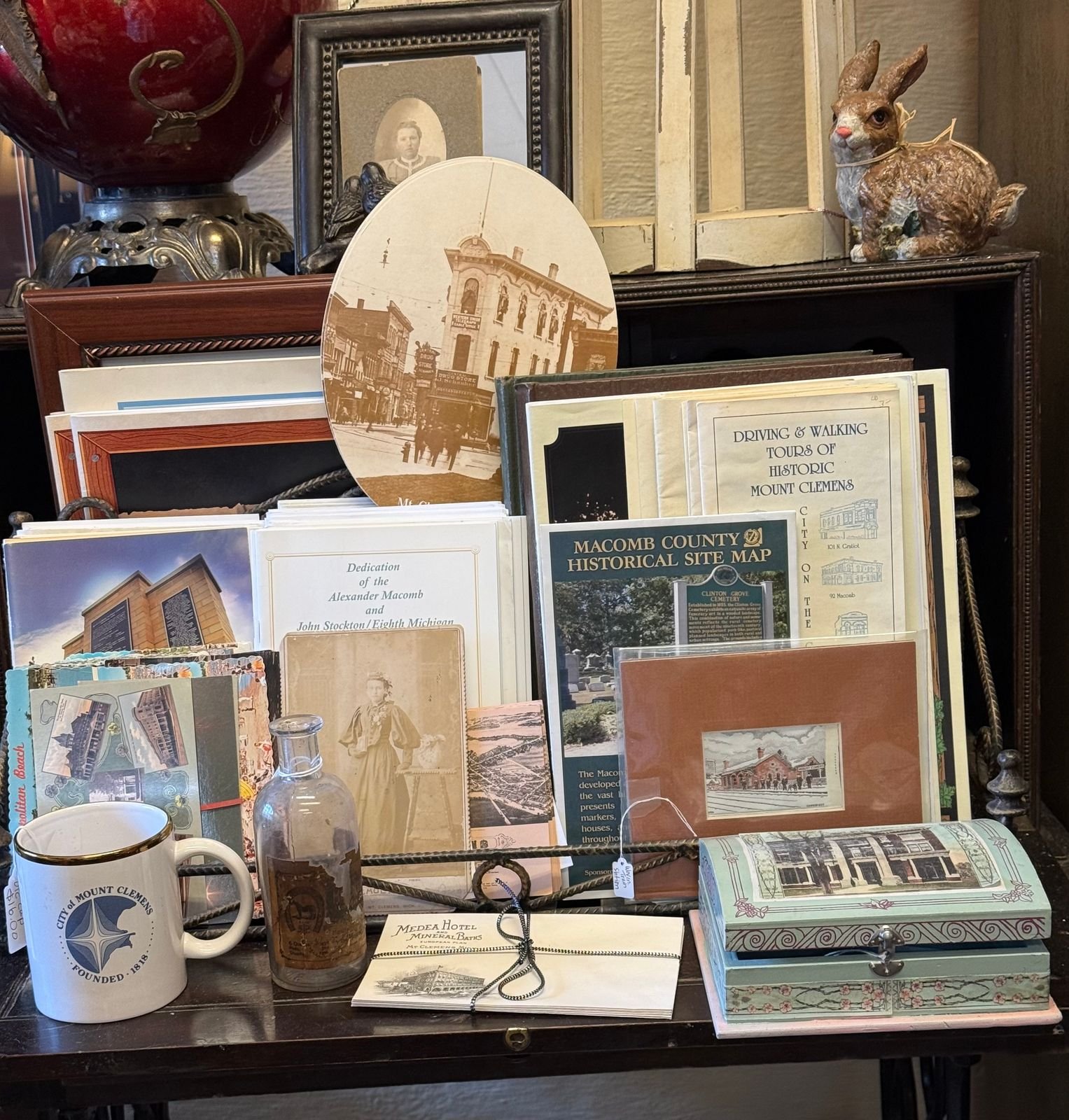 A table display of Mount Clemens memorabilia including a City of Mount Clemens mug marked Founded 1818, an embossed glass bottle, Medea Hotel and Mineral Baths stationery tied with cord, an oval sepia photograph of downtown, Victorian cabinet card portraits, a Macomb County Historical Site Map, historic walking tour booklets, postcards, and a framed watercolor of the old depot.