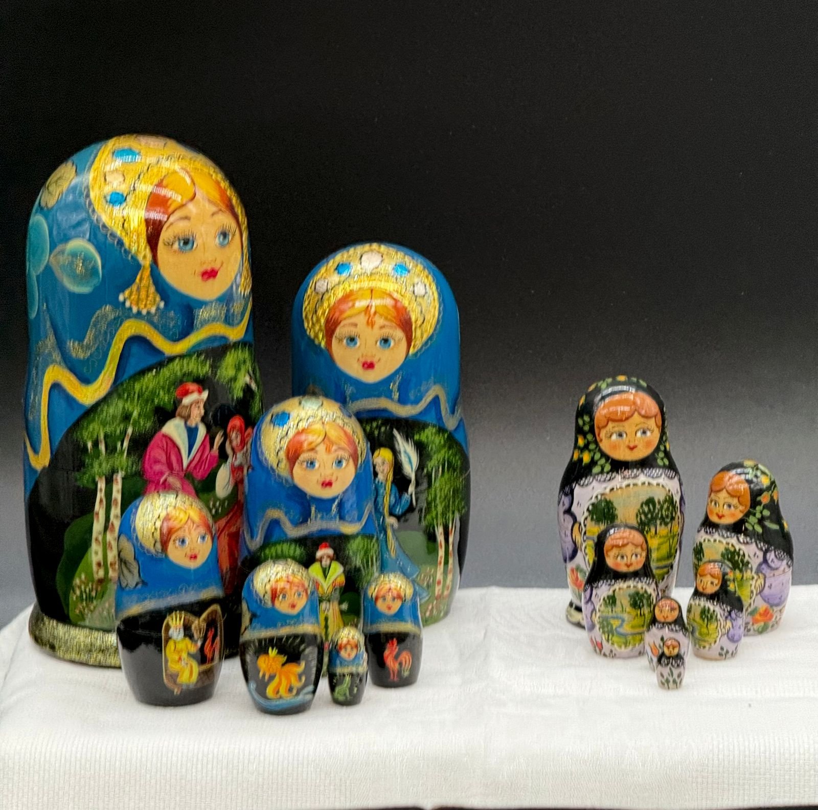 Two sets of hand painted Russian matryoshka nesting dolls arranged on white cloth. The seven-doll blue set wears gold kokoshnik headdresses with painted forest scenes and black lacquer panels showing a crowned tsar, a firebird, a golden fish, and a golden cockerel. The six-doll set beside it wears black floral headscarves with lavender aprons painted with green landscapes.