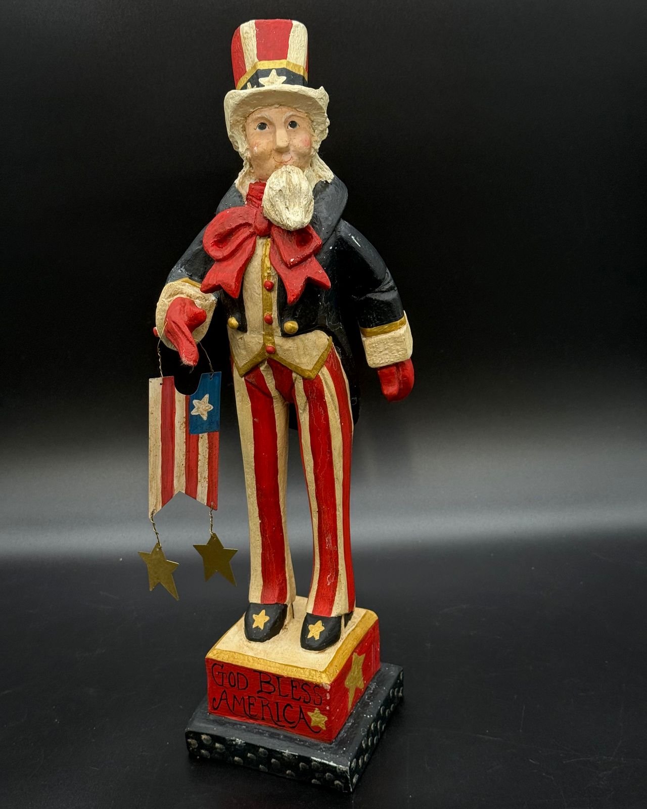 A folk art style House of Hatten Uncle Sam figure against a black backdrop. He wears a tall red and cream striped top hat with a gold band and white star, a white beard, a big red bow, a cream vest with gold buttons, a black tailcoat with gold-trimmed cuffs, red mittens, and red and cream striped trousers with star-topped black shoes. A small hand-painted flag hangs from one hand on twisted wire with two brass stars dangling below. He stands on a red pedestal lettered God Bless America with gold stars, set on a black dotted base.