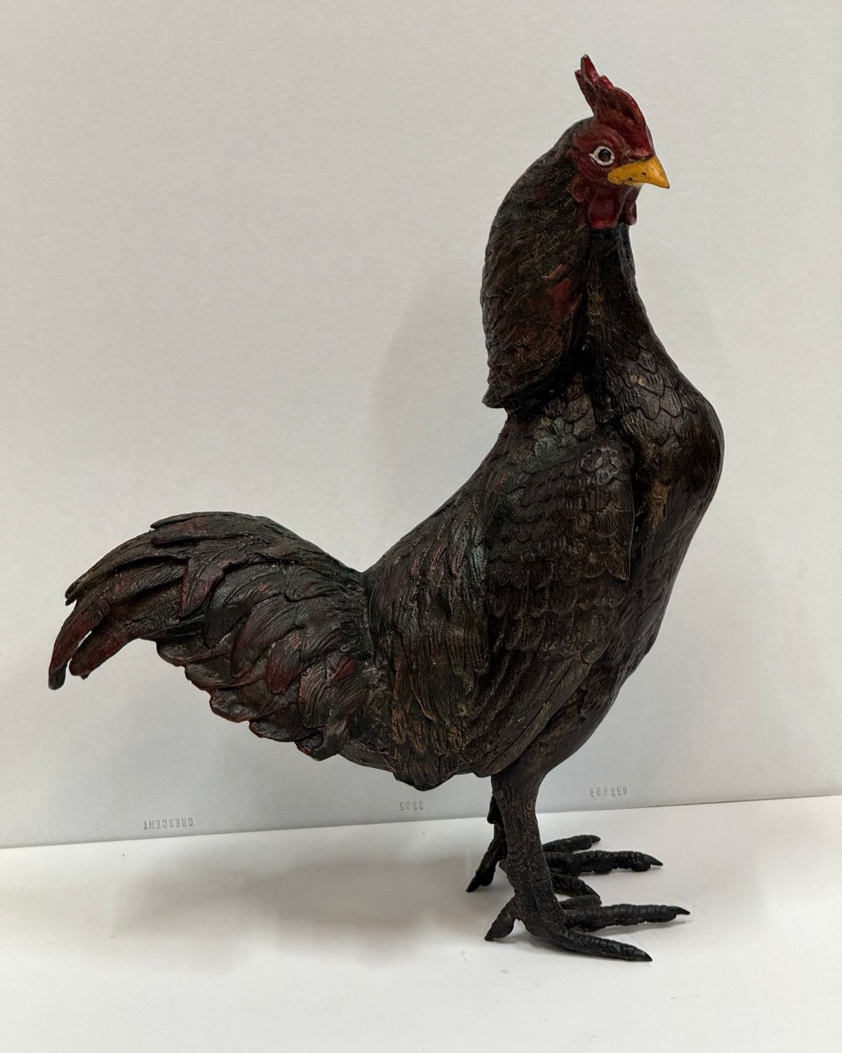 Bronze sculpture of a standing rooster with dark painted feathers, a red comb and wattles, and a yellow beak, on a white backdrop.