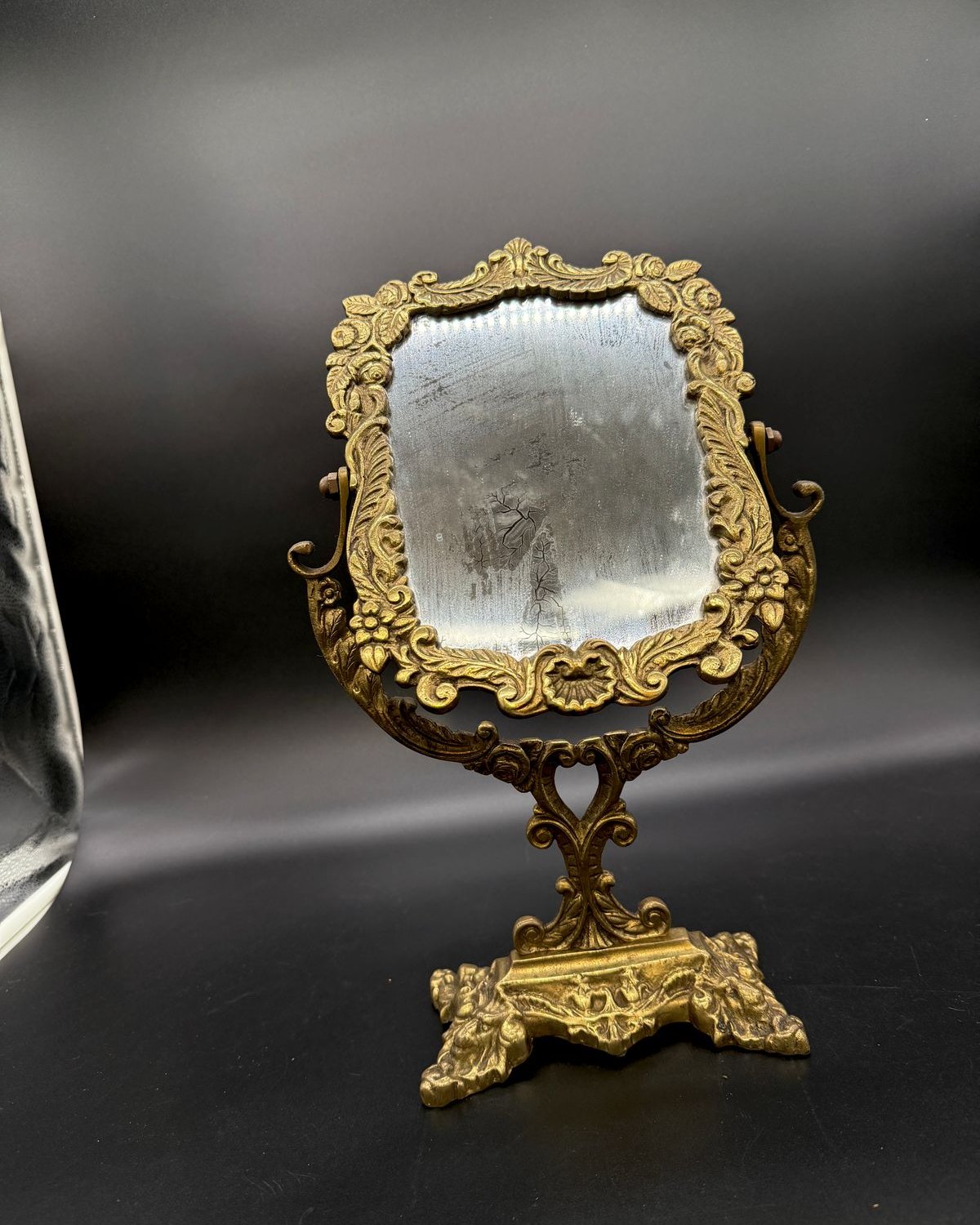 Vintage brass tabletop vanity mirror with an ornate cast frame of roses, leaves and scrolls, a swivel cradle, and a sculpted footed base, on a dark backdrop.