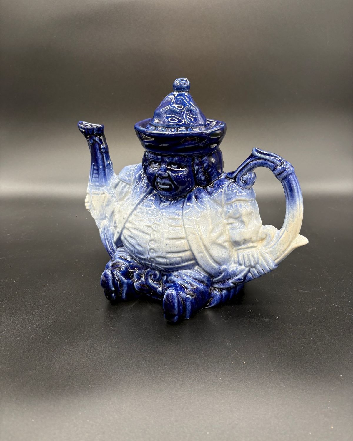 Blue and white flow blue figural teapot shaped as a seated robed man wearing a tasseled hat that forms the lid, with one arm as the spout and a curved handle, cobalt glaze flowing into cream.