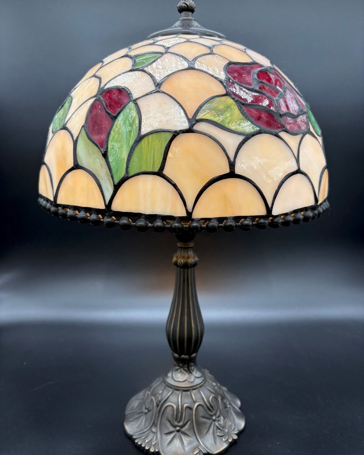 Vintage Tiffany-style table lamp with a leaded stained glass shade of red roses and green leaves on cream cathedral glass, beaded fringe along the bottom, on an ornate cast metal base.